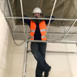 Pracownik w kasku i kamizelce odblaskowej stoi na drabinie, opierając się o konstrukcję sufitu podwieszanego w trakcie montażu.