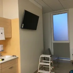 Telewizor LCD zamontowany na ścianie w pomieszczeniu medycznym, widoczna drabina, krzesła i umywalka z dozownikami mydła.