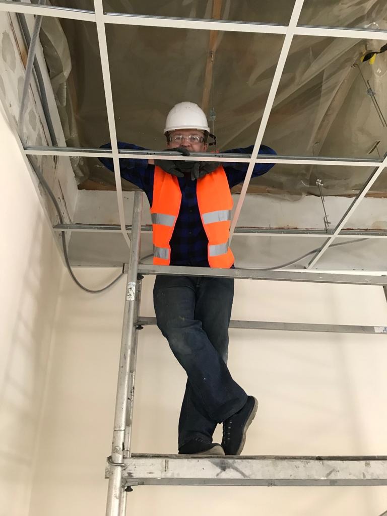 Pracownik w kasku i kamizelce odblaskowej stoi na drabinie, opierając się o konstrukcję sufitu podwieszanego w trakcie montażu.