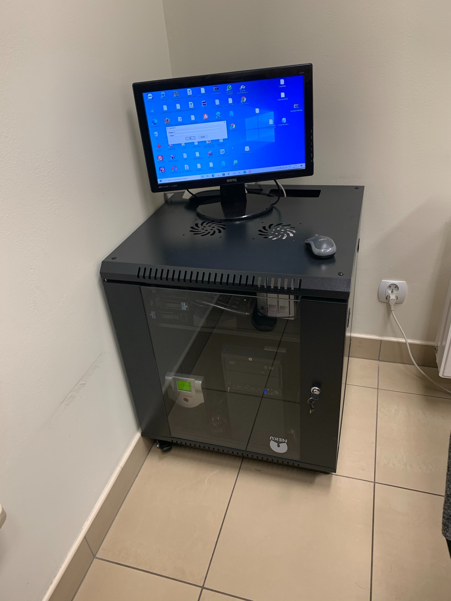 Czarna szafa serwerowa z zamontowanym monitorem BenQ, myszką Logitech, widoczne okablowanie i listwa zasilająca, na ekranie komputera otwarte okno logowania.