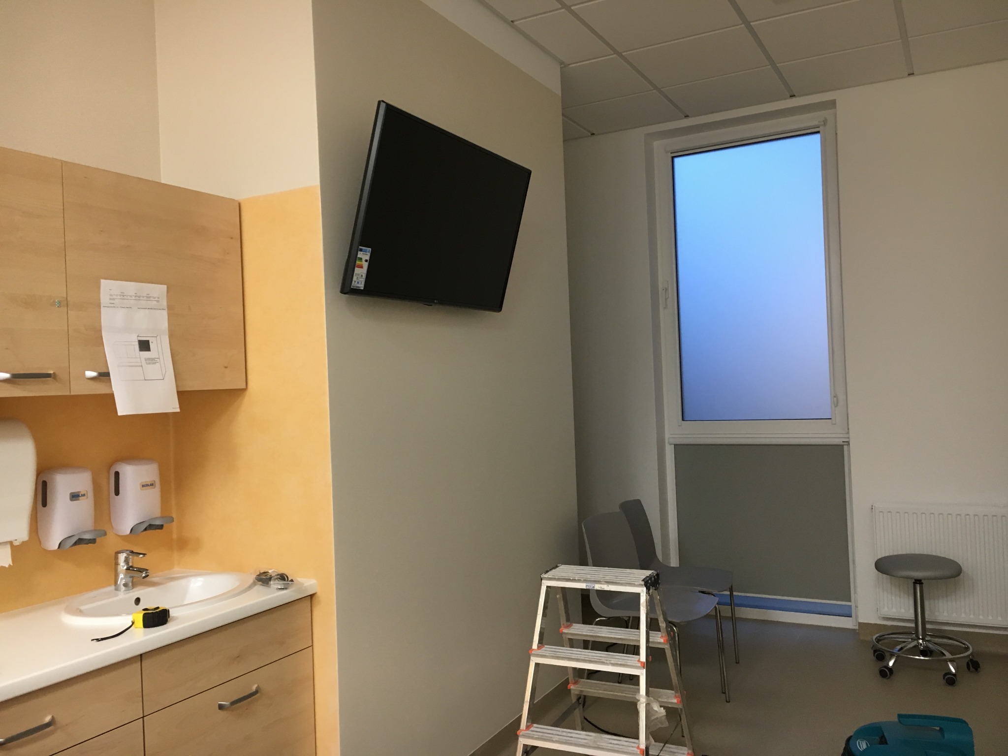 Telewizor LCD zamontowany na ścianie w pomieszczeniu medycznym, widoczna drabina, krzesła i umywalka z dozownikami mydła.