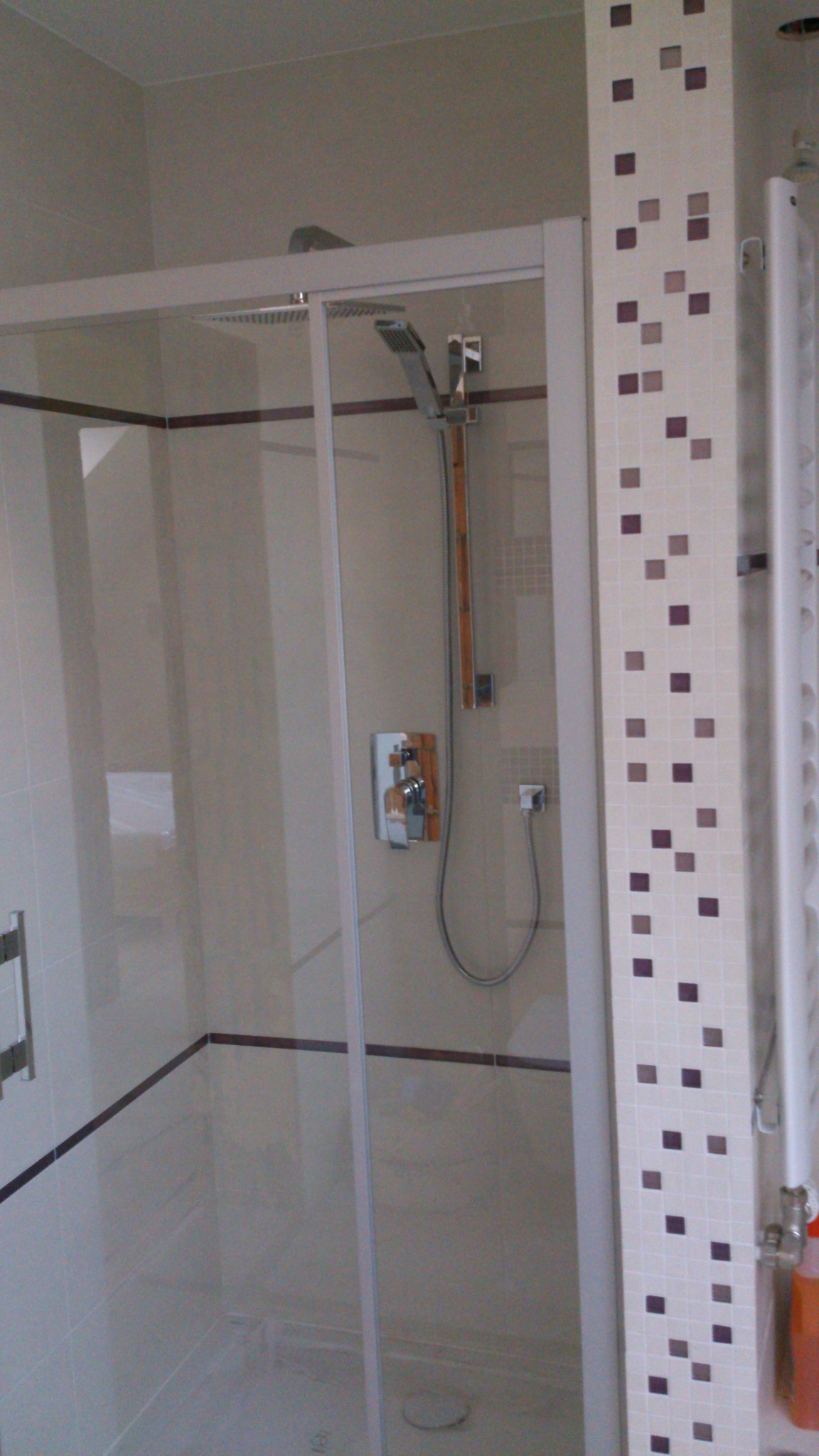 Kabina prysznicowa z przesuwanymi drzwiami, chromowaną armaturą i deszczownicą, obok pionowy grzejnik drabinkowy i dekoracyjny pasek z mozaiki w odcieniach fioletu.