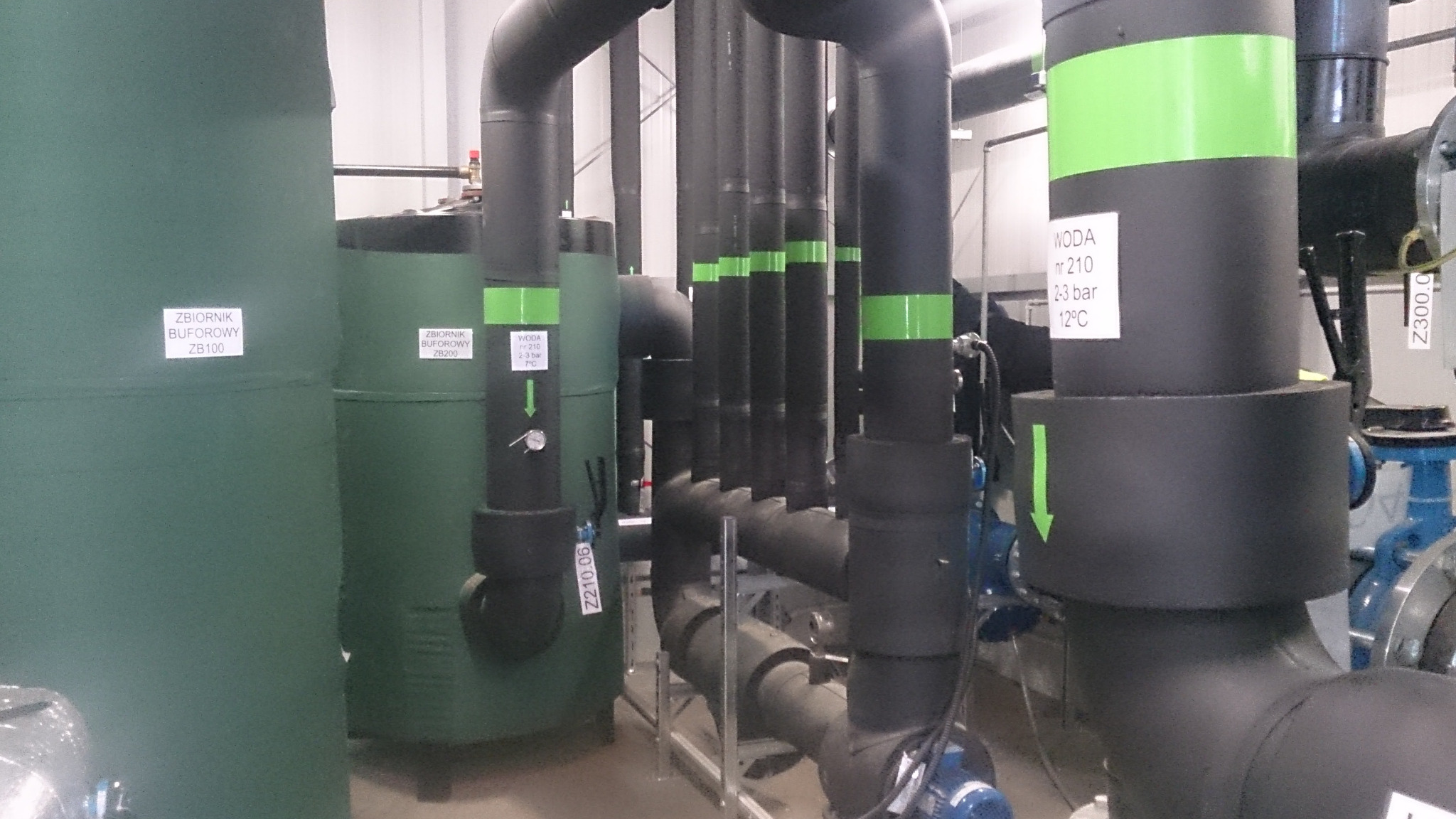 Instalacja kotłowni z zbiornikami buforowymi oznaczonymi ZB100 i ZB200, oraz siecią rur w izolacji z zielonymi oznaczeniami, opis parametrów wody na rurach.