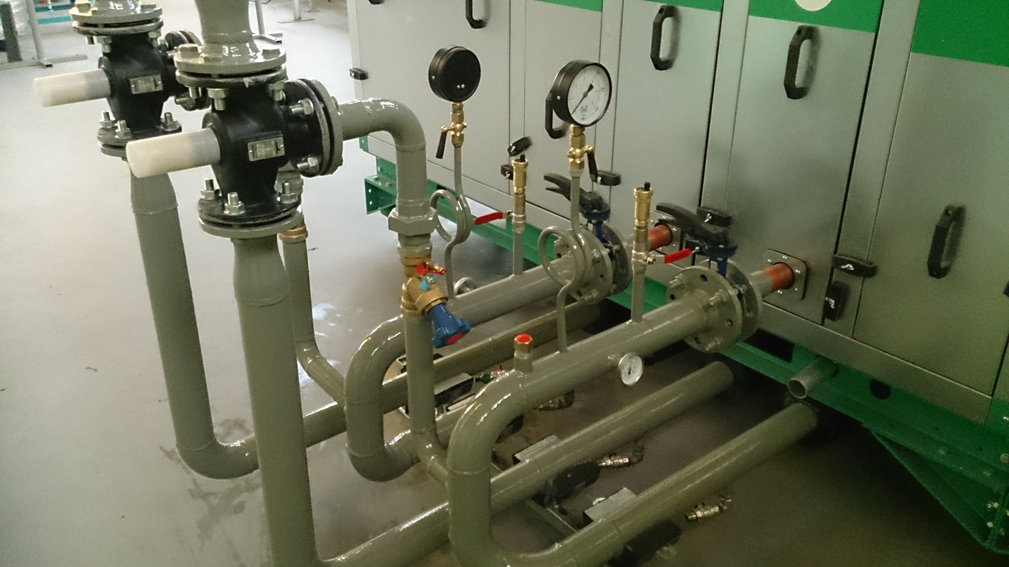 Szare rury instalacyjne z czarnymi manometrami i zaworami z czerwonymi i niebieskimi pokrętłami, podłączone do szarej obudowy urządzenia z zielonymi akcentami.