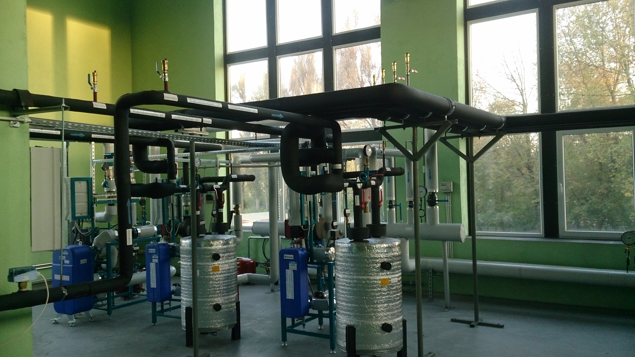 Kompleksowa instalacja grzewcza w kotłowni z rurami w izolacji, zbiornikami akumulacyjnymi w srebrnej folii i niebieskimi modułami, widok na zewnątrz przez okna.