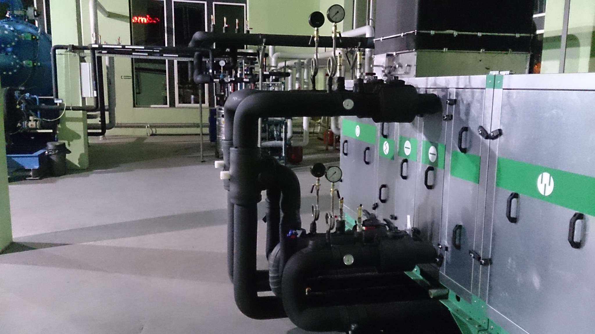 Kompleksowa instalacja przemysłowa z czarnymi rurami, manometrami i srebrną obudową z zielonymi akcentami w hali produkcyjnej.