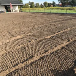 Przygotowany pod trawnik teren z charakterystycznymi śladami po bronowaniu, widoczny dom z tarasem i ogrodzenie w tle.