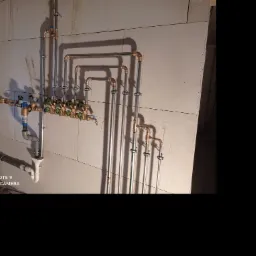 Rozbudowana instalacja hydrauliczna z rur miedzianych i PCV, zawory kulowe, manometr, zamocowana na ścianie z białych bloczków, widoczne cienie i ślady po montażu.