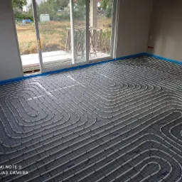 Białe rury ogrzewania podłogowego ułożone spiralnie na szarej macie izolacyjnej w surowym pomieszczeniu z dużym oknem balkonowym, widoczne niebieskie taśmy brzegowe.