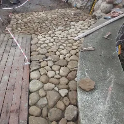 Niedokończona ścieżka z okrągłych kamieni przechodząca w drewniany taras, oddzielona taśmą ostrzegawczą, z betonowym murem po prawej stronie i kamiennym murem w tle.