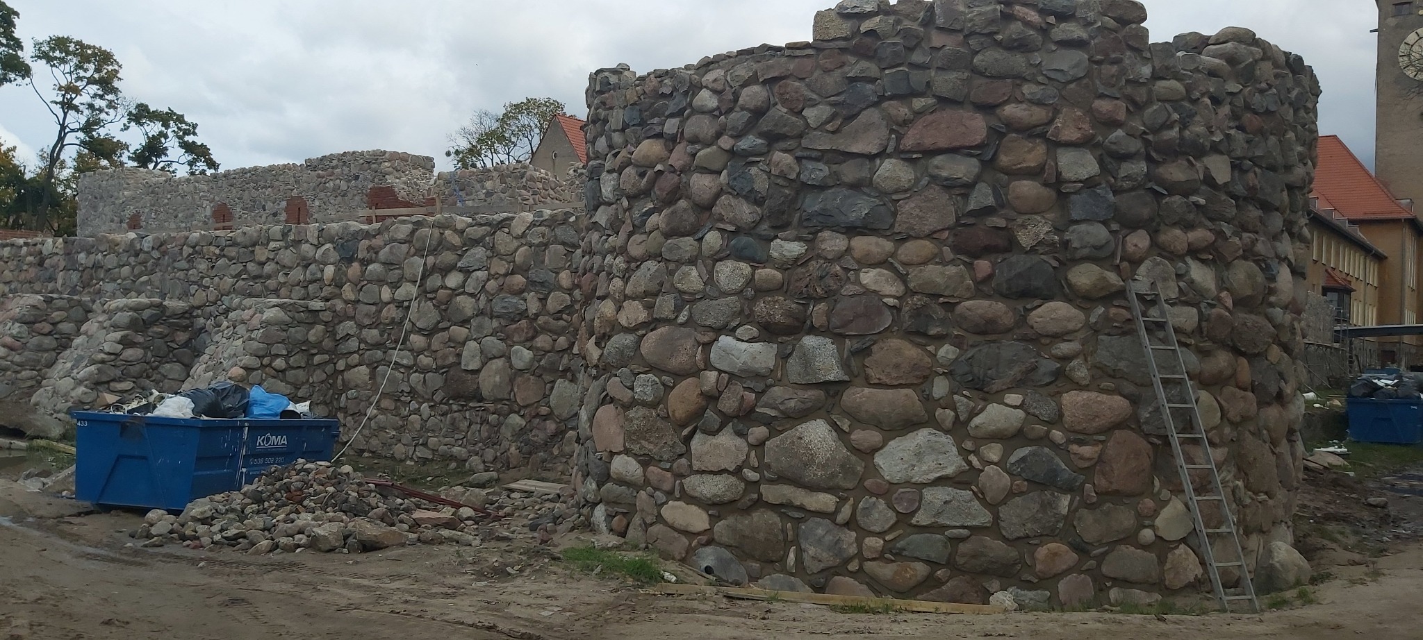Budowa kamiennego muru z okrągłą wieżą, widoczny kontener na gruz i drabina oparta o ścianę, w tle budynek z zegarem.