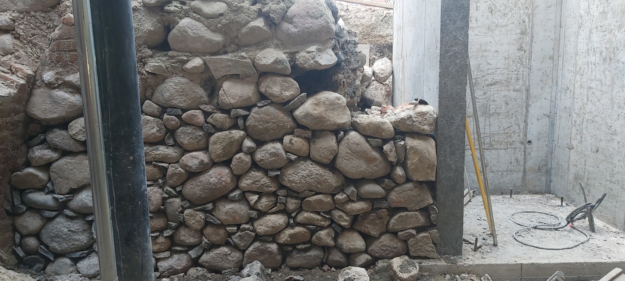 Kamienna ściana z nieregularnych, szarych kamieni, częściowo wykończona, z widocznymi fragmentami cegieł w tle i elementami konstrukcji betonowej po prawej stronie.