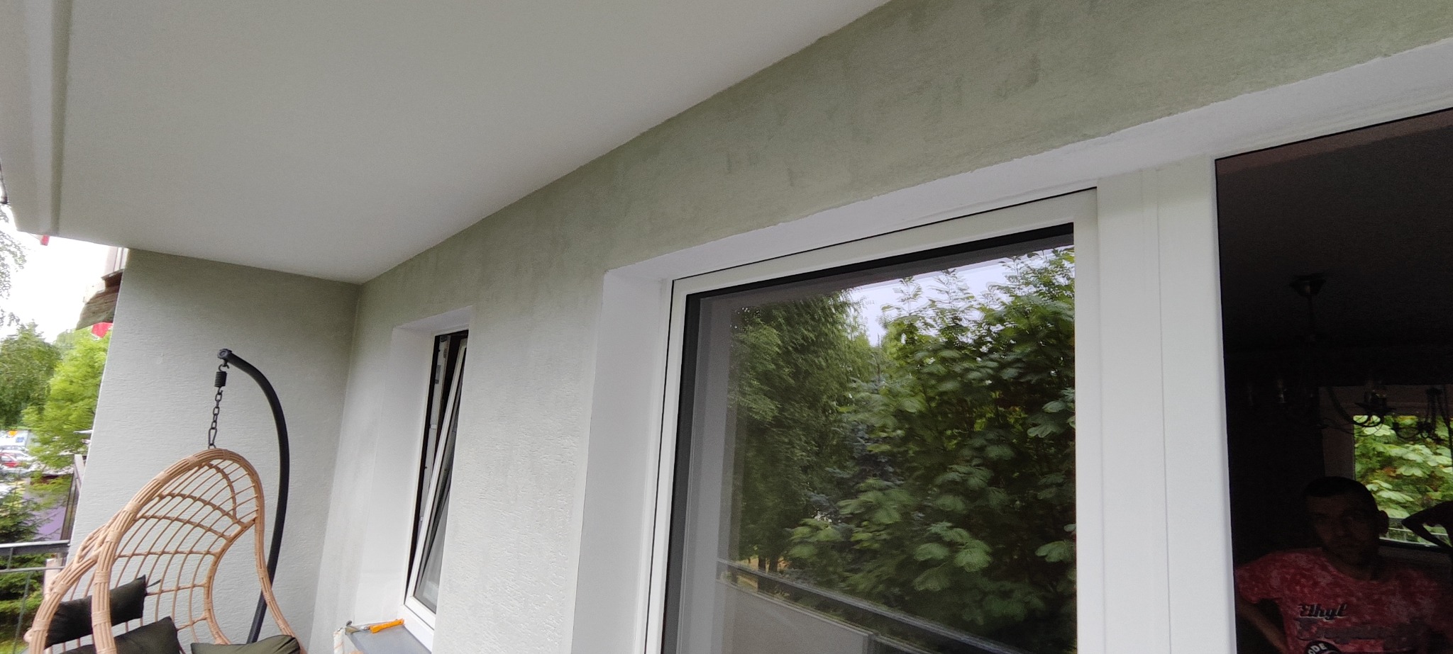 Wykończony balkon z oknami PCV i wiszącym fotelem. Ściany w odcieniach szarości, widok na zieleń za oknem. Minimalistyczny design.