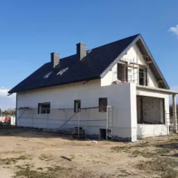 Dom jednorodzinny w trakcie prac elewacyjnych, widoczne rusztowania, częściowo otynkowane ściany i niezabudowany taras.