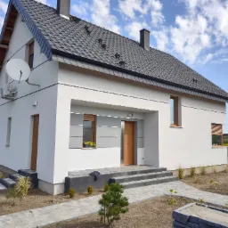 Nowoczesny dom jednorodzinny z jasną elewacją, grafitowym dachem, schodami wejściowymi wyłożonymi kostką i zagospodarowanym terenem wokół