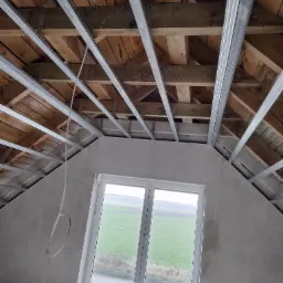 Poddasze w trakcie adaptacji: widok na drewnianą konstrukcję dachu z zamontowanymi metalowymi profilami pod płyty gipsowo-kartonowe, surowa ściana szczytowa z oknem na tle zielonego pola.