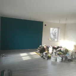 Pokój w trakcie malowania: jedna ściana pomalowana na turkusowo, reszta biała, podłoga szara, widoczne puszki z farbą i narzędzia malarskie.