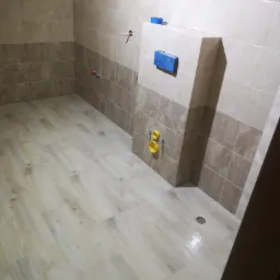 Wnętrze łazienki w trakcie remontu; widoczne płytki na ścianach i podłodze imitujące drewno, zabudowa stelaża podtynkowego WC z niebieskimi elementami i żółtymi rurami.