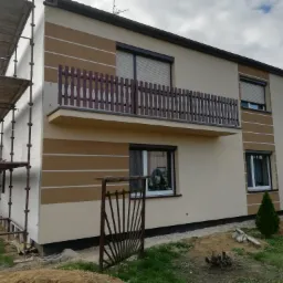 Dwukondygnacyjny dom z ocieploną elewacją w beżowo-brązowe pasy, balkon z drewnianą balustradą, rusztowanie przy bocznej ścianie, teren wokół domu w trakcie prac wykończeniowych.