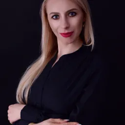 Portret uśmiechniętej blondynki w czarnej koszuli z założonymi rękami i złotym zegarkiem na czarnym tle.