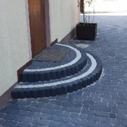 Wykończone wejście do domu z trzema półkolistymi stopniami z kostki brukowej w kolorze grafitowym i białym, prowadzące do drewnianych drzwi z wycieraczką. Podłoże wyłożone grafitową kostką brukową.
