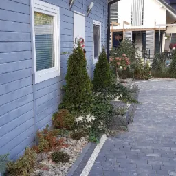 Szary dom z drewnianą elewacją, oświetleniem zewnętrznym i brukowanym podjazdem, ozdobiony roślinami wzdłuż ściany i w donicach.