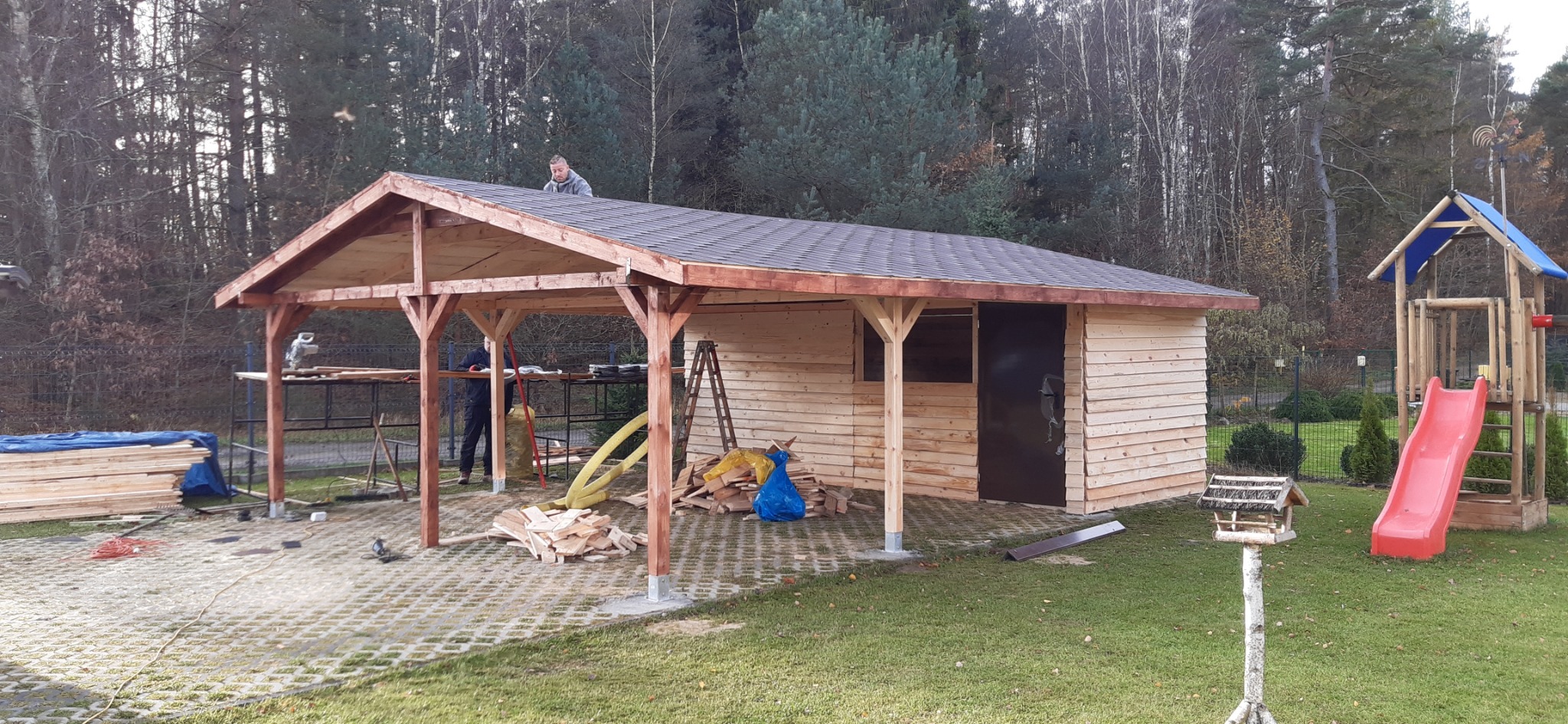Budowa drewnianej altany z zadaszeniem z ciemnej dachówki bitumicznej w ogrodzie, widoczne elementy konstrukcyjne, narzędzia pracy, oraz plac zabaw w tle.