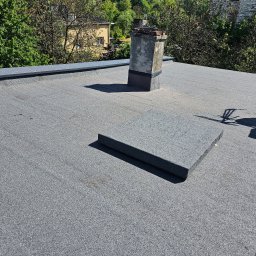 Sunlit Energy - Szary, płaski dach pokryty papą z wystającym kominem i elementem wentylacyjnym, otoczony zielenią drzew i budynkami.