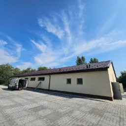 Budynek z nowym poszyciem z blachodachówki w kolorze brązowym, widoczny z dużej odległości na tle błękitnego nieba z chmurami, na placu manewrowym stoi bus z otwartymi drzwiami