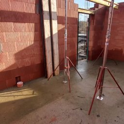 Sunlit Energy - Budowa domu z czerwonej cegły: ściany w trakcie wznoszenia, drewniane belki stropowe i metalowe podpory. Widoczne zbrojenia i mokra posadzka.