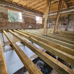 Sunlit Energy - Wnętrze budynku w trakcie remontu: drewniane belki stropowe, fragmenty muru z cegły i pustaków, okna. Widoczne narzędzia i materiały budowlane. Prace remontowe w toku.