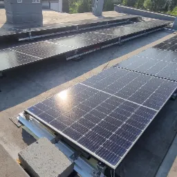 Rząd paneli słonecznych zamontowanych na płaskim dachu budynku, widoczne elementy konstrukcyjne i mocowania, z odbiciem słońca na powierzchni paneli.