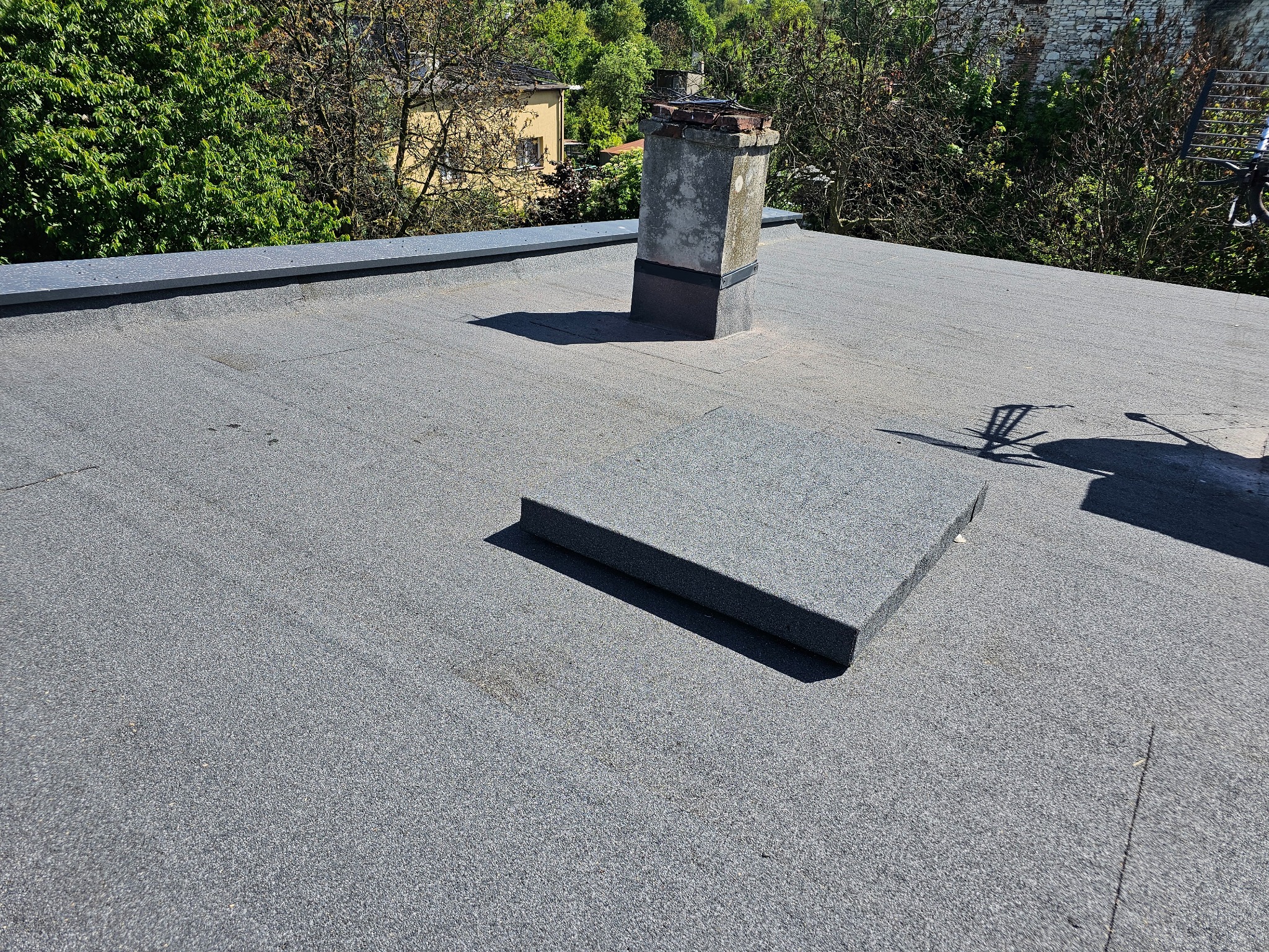 Szary, płaski dach pokryty papą z wystającym kominem i elementem wentylacyjnym, otoczony zielenią drzew i budynkami.