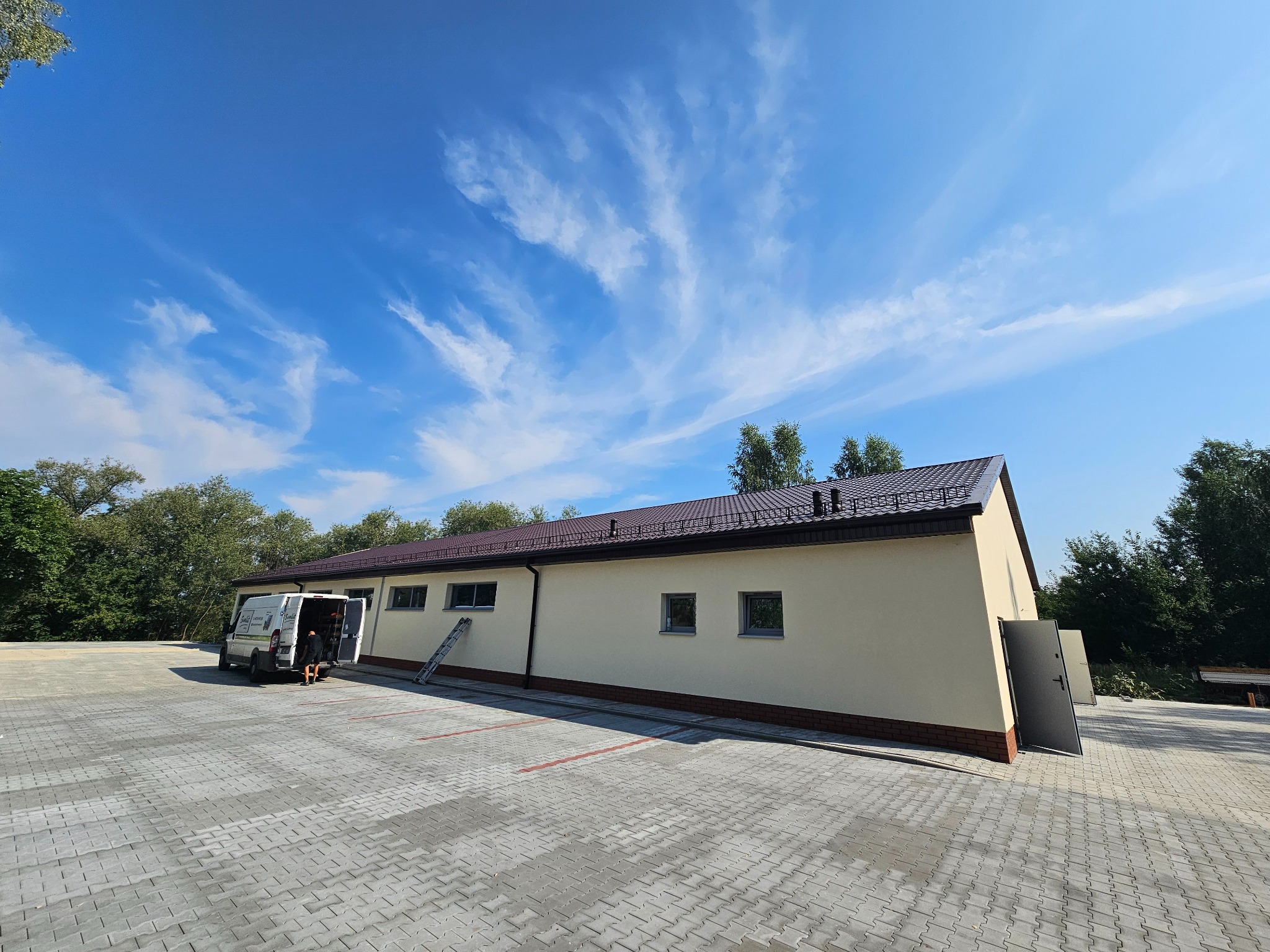 Budynek z nowym poszyciem z blachodachówki w kolorze brązowym, widoczny z dużej odległości na tle błękitnego nieba z chmurami, na placu manewrowym stoi bus z otwartymi drzwiami