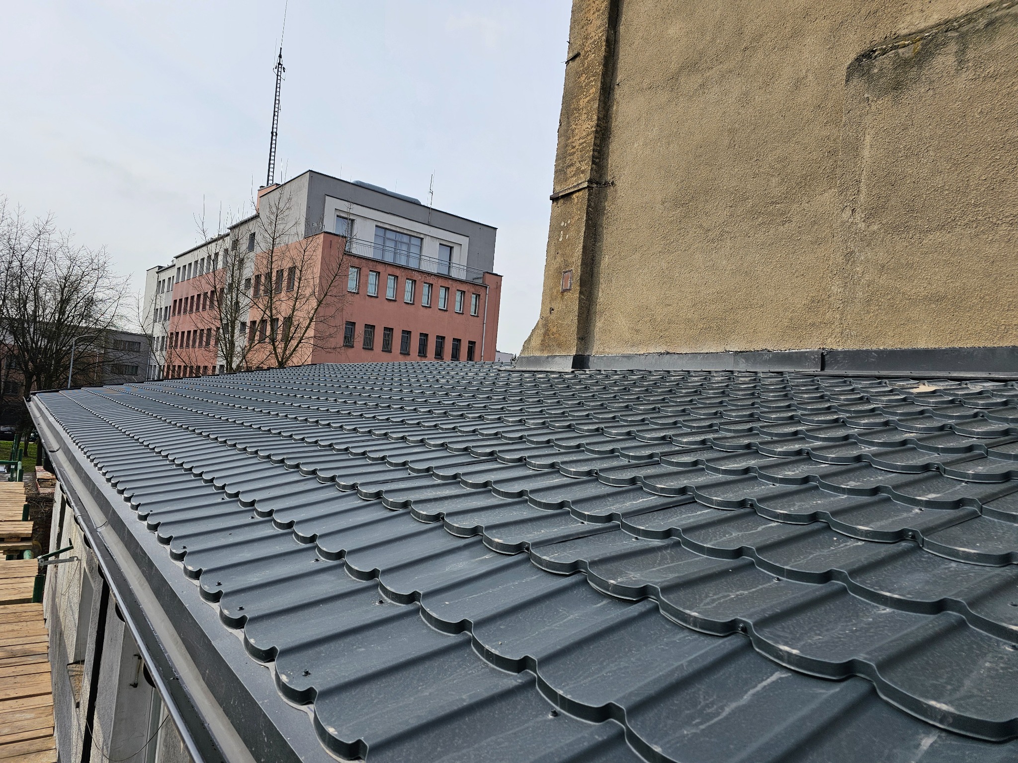Nowy, grafitowy dach z blachodachówki modułowej, widok z góry, z fragmentem rusztowania i budynkami w tle.