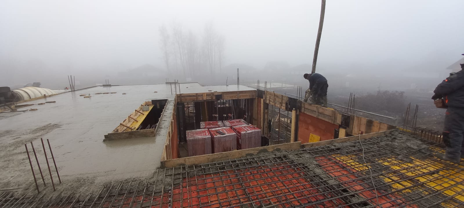 Wylewanie betonu na strop budynku w trakcie budowy, widoczne zbrojenia i szalunki. Prace budowlane w mglisty dzień.
