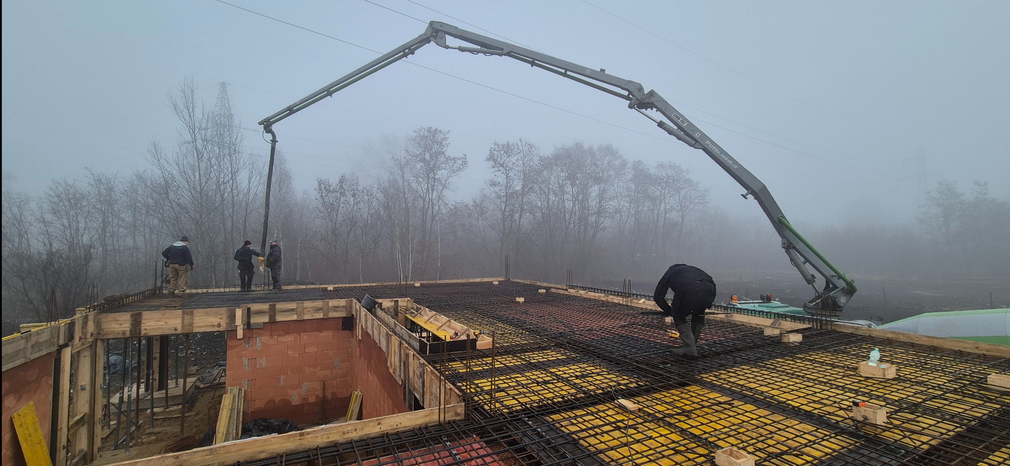 Betonowanie stropu budynku w trakcie budowy. Widoczna pompa do betonu, zbrojenie i pracownicy w odzieży roboczej. Mglista pogoda w tle.
