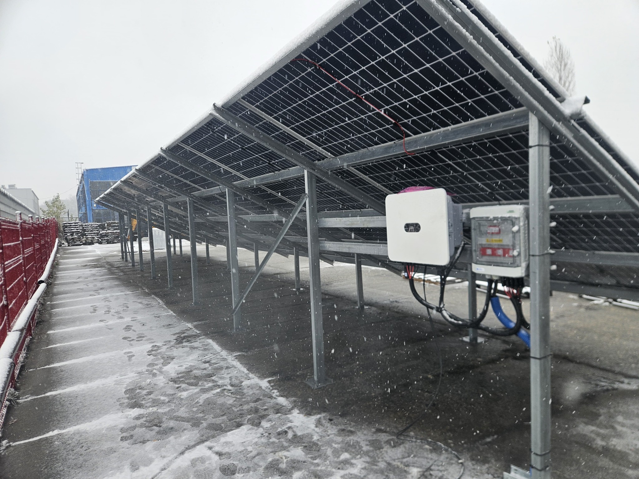 Instalacja paneli słonecznych pokryta śniegiem, widoczne inwertery i okablowanie. Śnieżny krajobraz w tle, metalowa konstrukcja wsporcza.