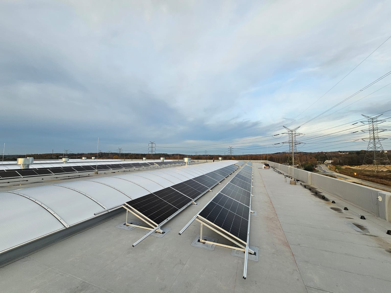 Rząd paneli słonecznych na płaskim dachu budynku przemysłowego, widok z góry, pochmurne niebo w tle, słupy wysokiego napięcia w oddali.