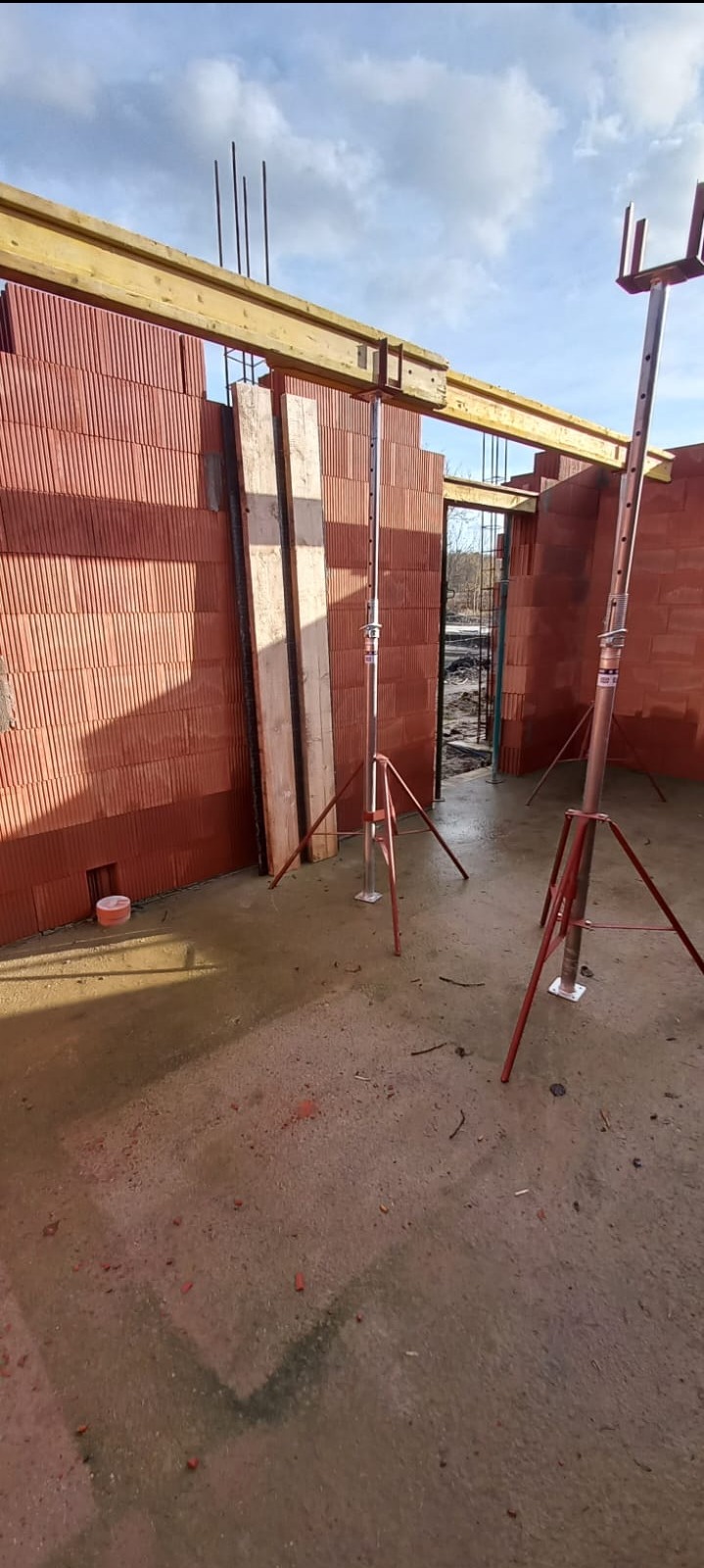 Budowa domu z czerwonej cegły: ściany w trakcie wznoszenia, drewniane belki stropowe i metalowe podpory. Widoczne zbrojenia i mokra posadzka.