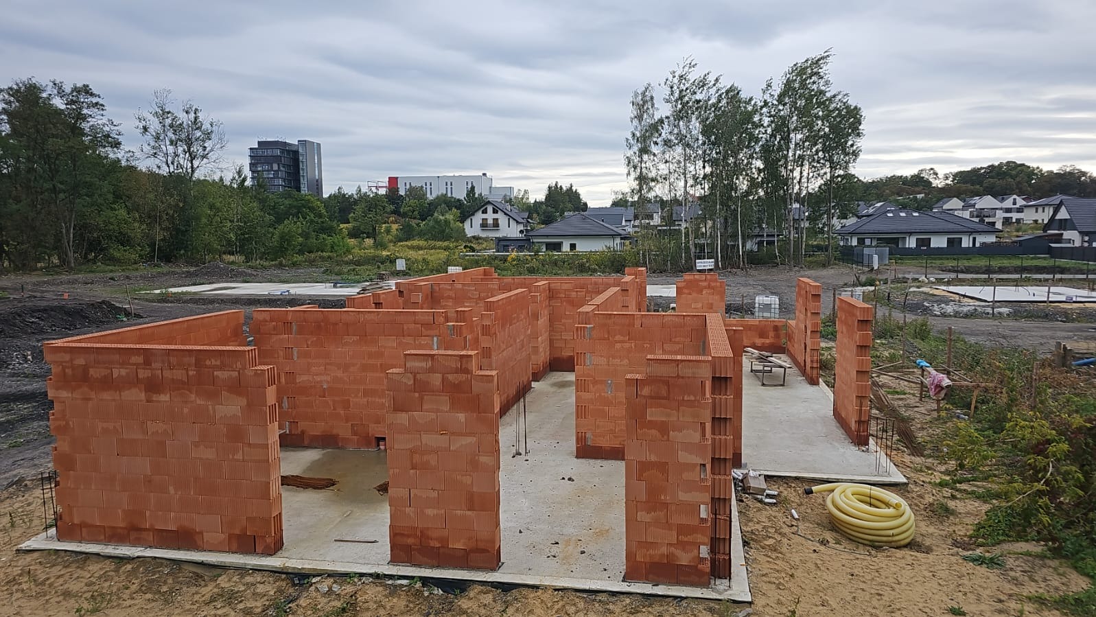 Budowa domu jednorodzinnego w trakcie wznoszenia ścian z czerwonej cegły. Widoczne fundamenty, zbrojenia i elementy instalacji. W tle zabudowa osiedlowa i zieleń.