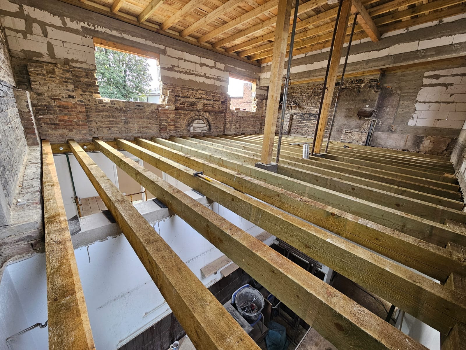 Wnętrze budynku w trakcie remontu: drewniane belki stropowe, fragmenty muru z cegły i pustaków, okna. Widoczne narzędzia i materiały budowlane. Prace remontowe w toku.