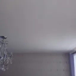 Kryształowy żyrandol zwisający z gładkiego, białego sufitu, na tle ściany pokrytej tapetą w subtelny, geometryczny wzór w odcieniach szarości, z fragmentem okna z fioletową zasłoną po prawej stronie.