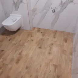 Nowoczesna łazienka z marmurowymi płytkami ściennymi i podłogą imitującą drewno, z wiszącą miską WC i chromowaną armaturą.