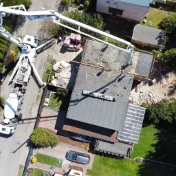 Widok z drona na budowę domu: beton pompowany z betonomieszarki na zbrojony fundament, obok stos cegieł i materiałów budowlanych.