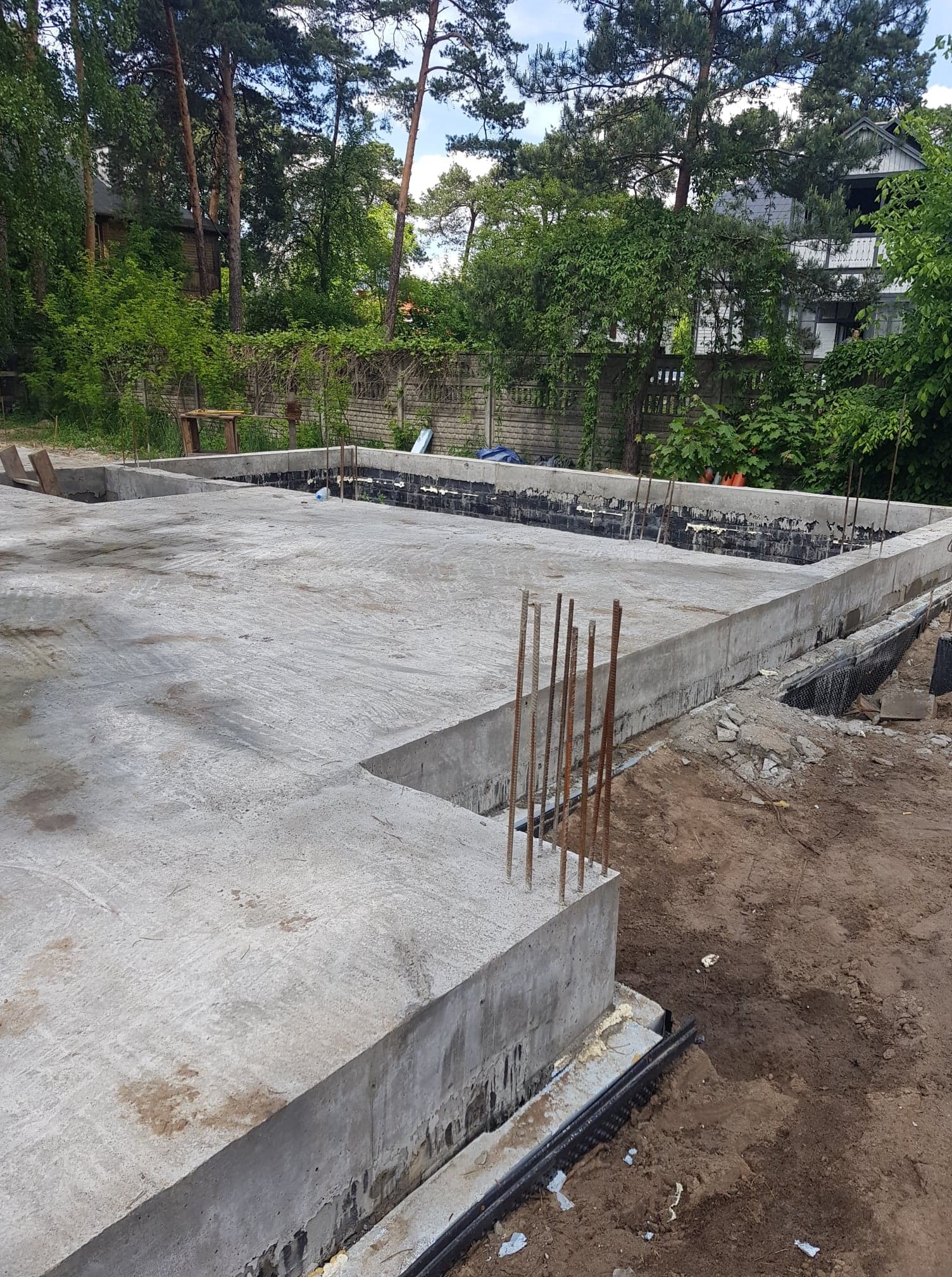 Wylany betonowy fundament domu z wystającymi prętami zbrojeniowymi, widoczne zabezpieczenia hydroizolacyjne, w tle drzewa i częściowo widoczny budynek.