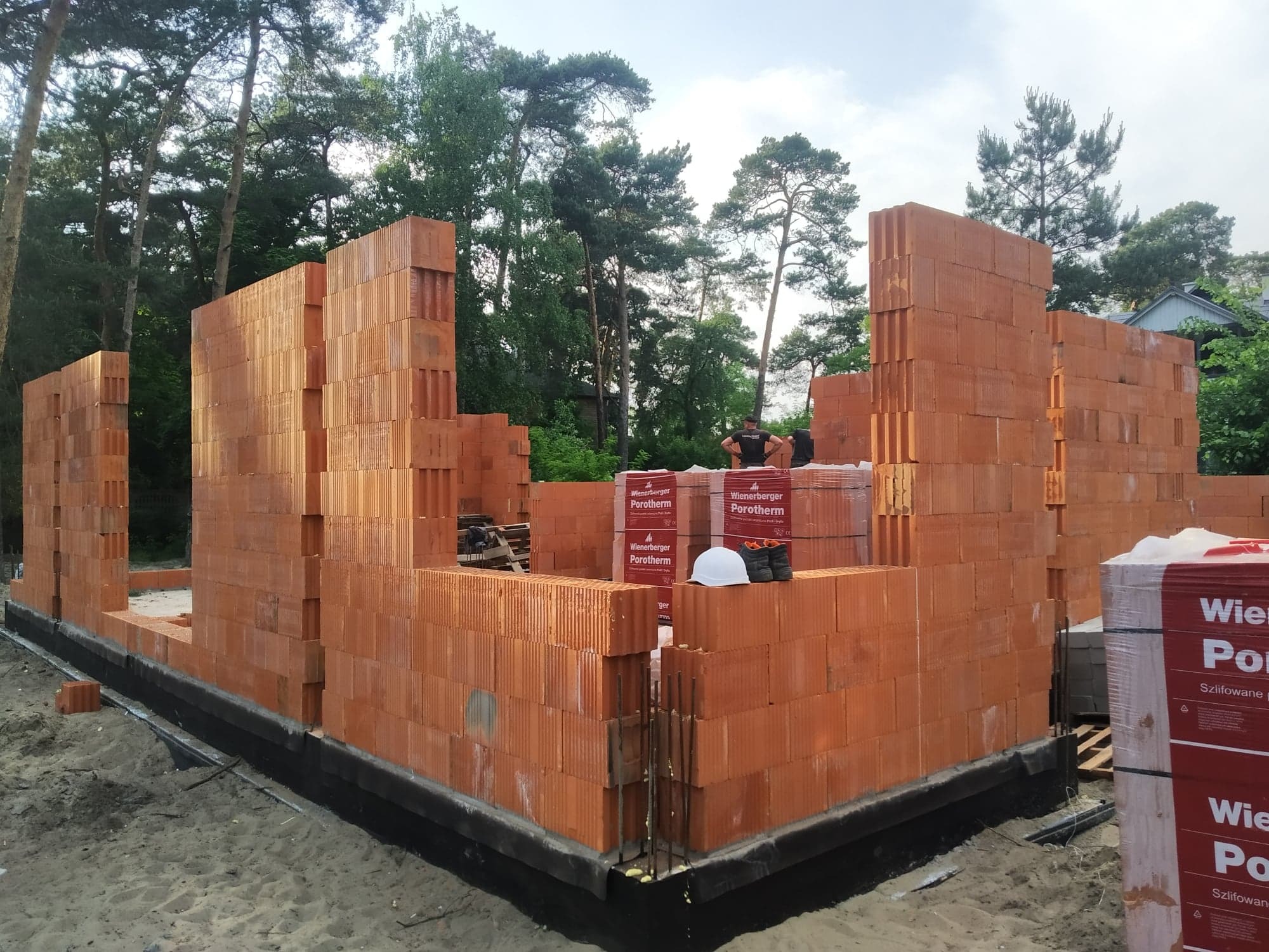 Wznoszenie ścian parteru z czerwonej ceramiki poryzowanej, widoczne palety z pustakami, biały kask i pracownik na budowie, w tle drzewa.