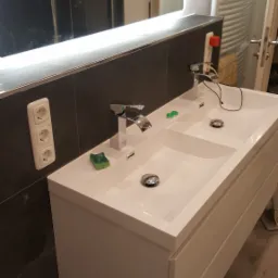 Nowoczesna, podwójna umywalka z białego konglomeratu z chromowanymi bateriami, zielonymi akcentami i podświetlanym lustrem w łazience z ciemnymi płytkami.