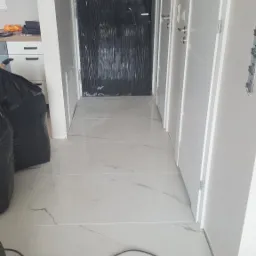 Białe marmuropodobne płytki podłogowe w korytarzu podczas remontu, drzwi zabezpieczone folią, widoczne worki z odpadami budowlanymi i kabel elektryczny na podłodze.