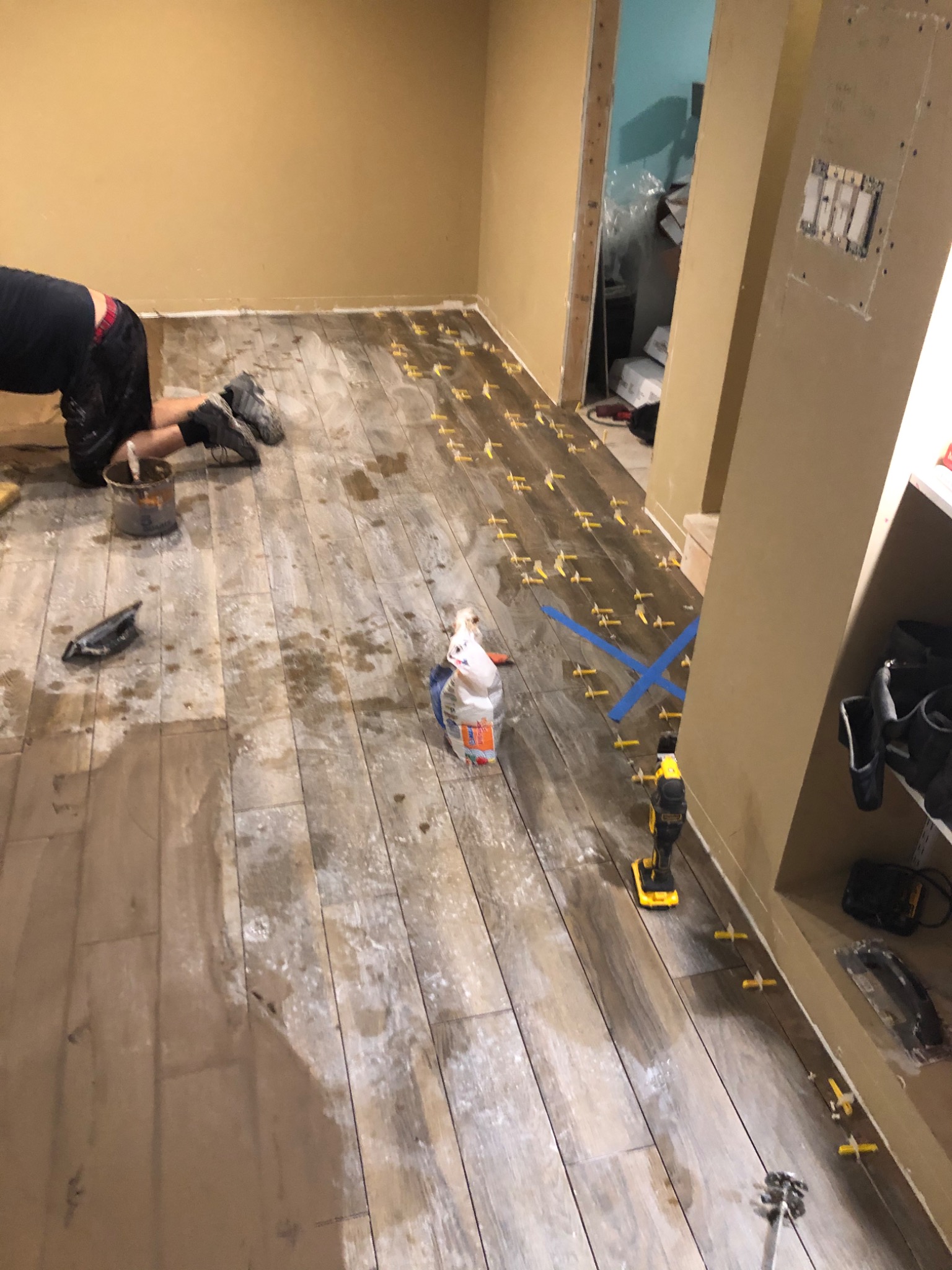 Układanie podłogi z płytek imitujących drewno, widoczne krzyżyki dystansowe, narzędzia i klej, w tle ściana przygotowana do montażu osprzętu elektrycznego.